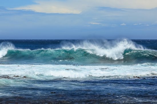 Waves breaking with foam under a blue sky with calm blue water closer to shore