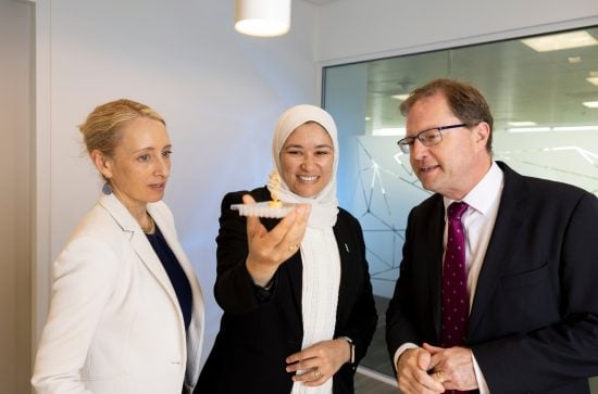 Woman in white jacket, woman in black jacket and white head cover holding a petri dish watched by a man in a black jacket
