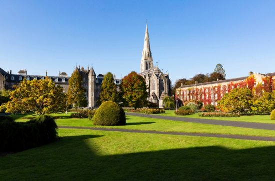 Church spire surrounded by creeper-clad buildings with green planted area in front and a blue sky above